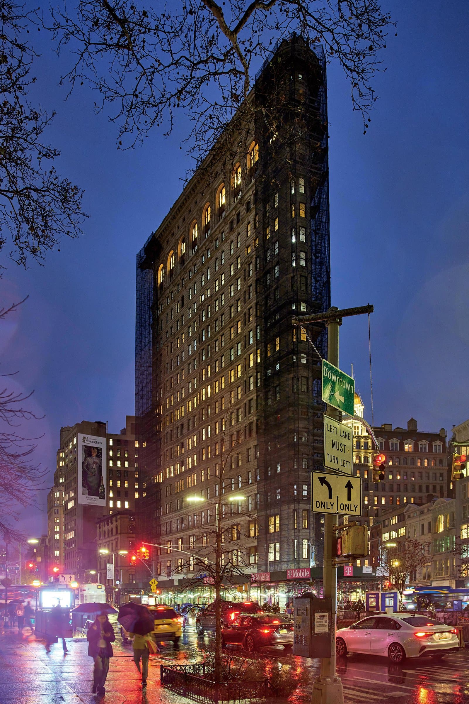 Tall narrow building above wet streets, cars and people with umbrellas